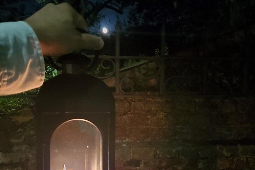 Una mano sosteniendo un farol con llama encendida, sobre un fondo oscuro que muestra un muro de ladrillo y una barandilla metálica. 2