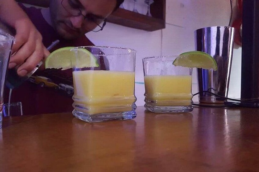 Two glasses of a yellow beverage with lime wedges, a bar tool, and a shaker on a wooden surface. 2
