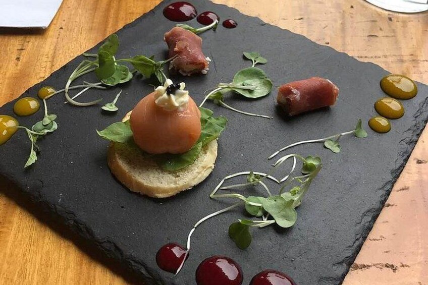 A slate board with a piece of bread topped with a round meat item, garnished with greens and olives. 1