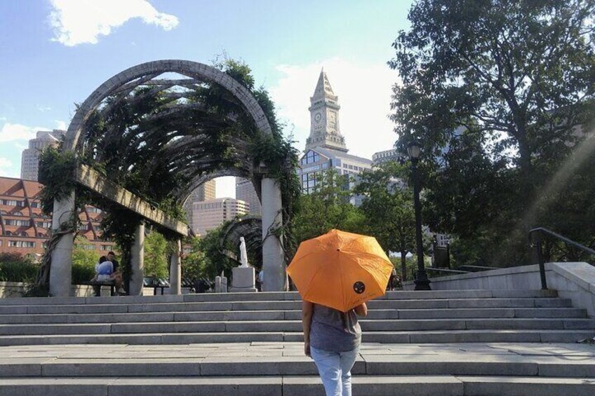 A person walking up steps with an umbrella, a stone archway, and a clock tower in the background. 4