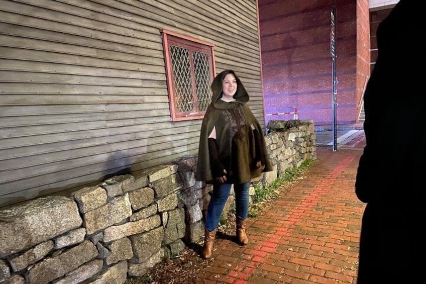 A person standing next to a stone wall with a metal window, wearing a dark coat and boots. 4