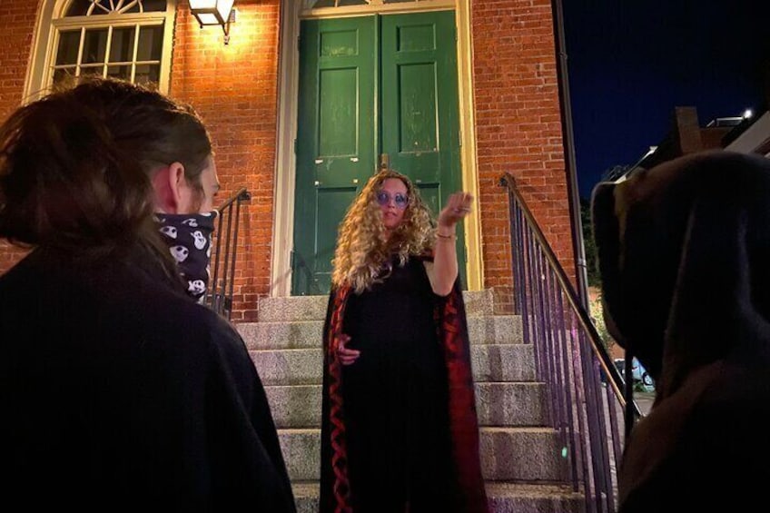 A person in a black dress with a red cape standing on a staircase in front of a brick building with a green door. 1