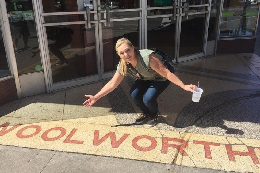 En person som hukar på en trottoar och håller en kopp i handen, framför en butik med en skylt där det står ”WOOLWORTH”. 2