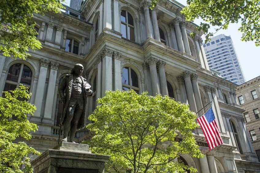 A statue in front of a neoclassical building with columns, an American flag, and a tree. 3
