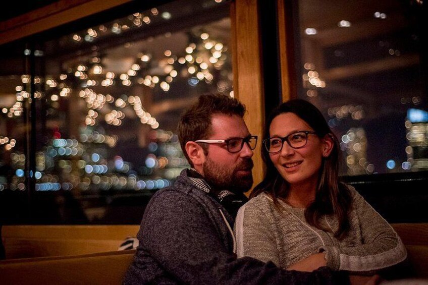 Two people sitting close together in a dimly lit room with a cityscape visible through a window. 2