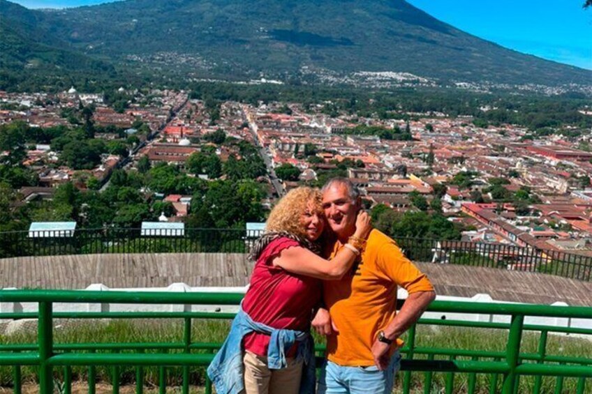 Una pareja se abraza en una baranda verde, con un paisaje urbano y montañas al fondo. 4