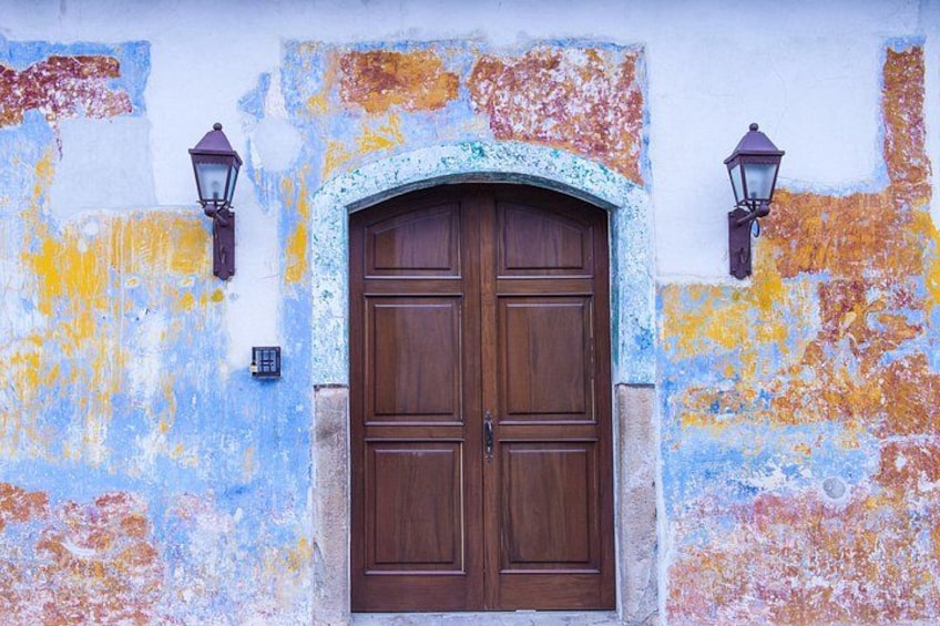 Une façade de bâtiment usée par les intempéries, avec une porte à double battant en bois, deux lanternes fixées au mur et une peinture colorée, dont les couches s'écaillent. 4