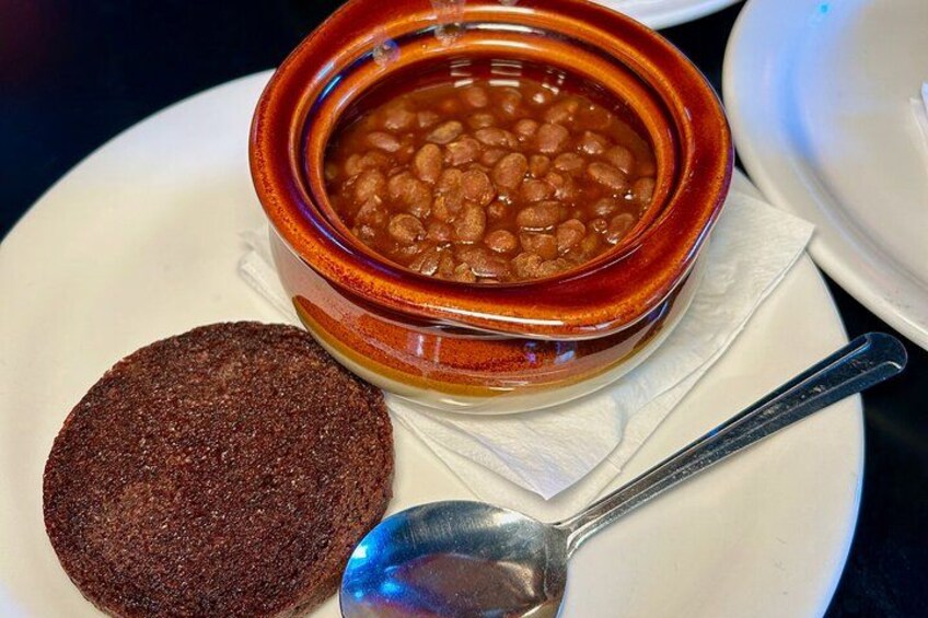 A bowl of baked beans, a slice of dark bread, and a spoon on a plate. 4