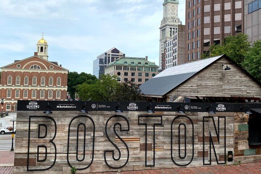 A wooden sign with 'BOSTON' in large letters, a historic building, and a modern skyscraper in the background. 2