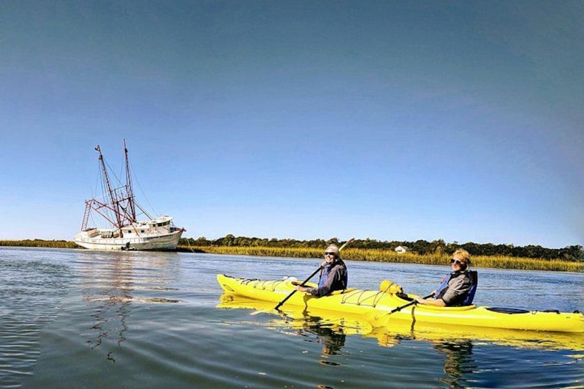Dos personas remando en kayak por un río tranquilo con un velero al fondo. 2