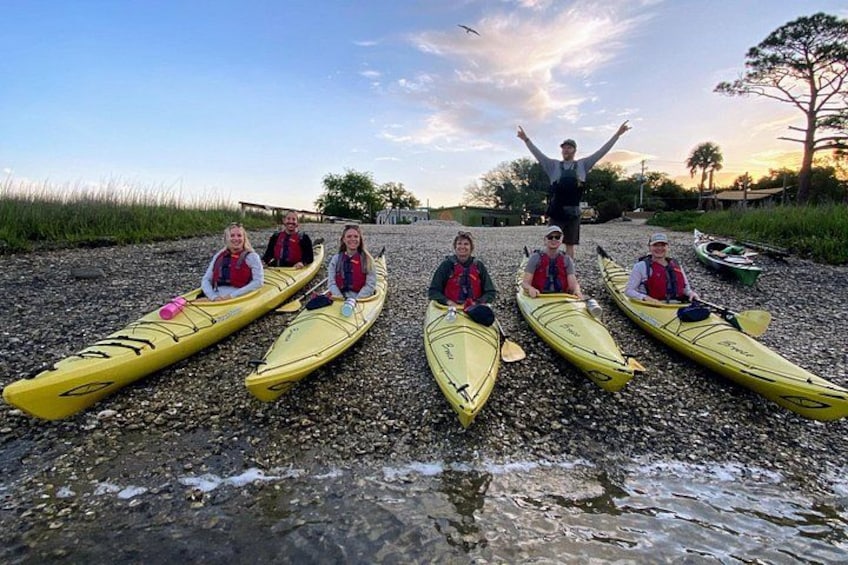 A group of people in kayaks on a pebbled shore with a person standing in the background. 2