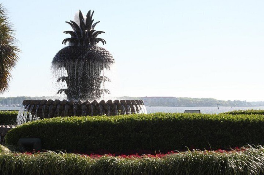 A fountain with water jets, surrounded by neatly trimmed hedges and red flowers, with a body of water and distant buildings in the background. 5