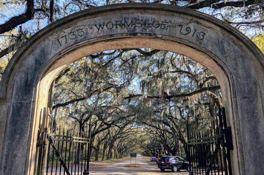 An arched stone gateway with the inscription '1733 Wormsloe 1913' and a gated pathway leading through a tree-lined avenue. 2