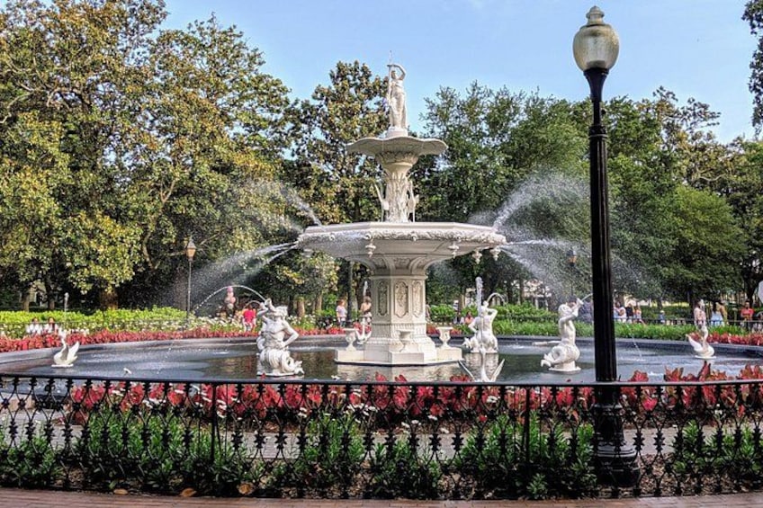 A fountain with multiple water jets, surrounded by a garden with red flowers and trees. 1