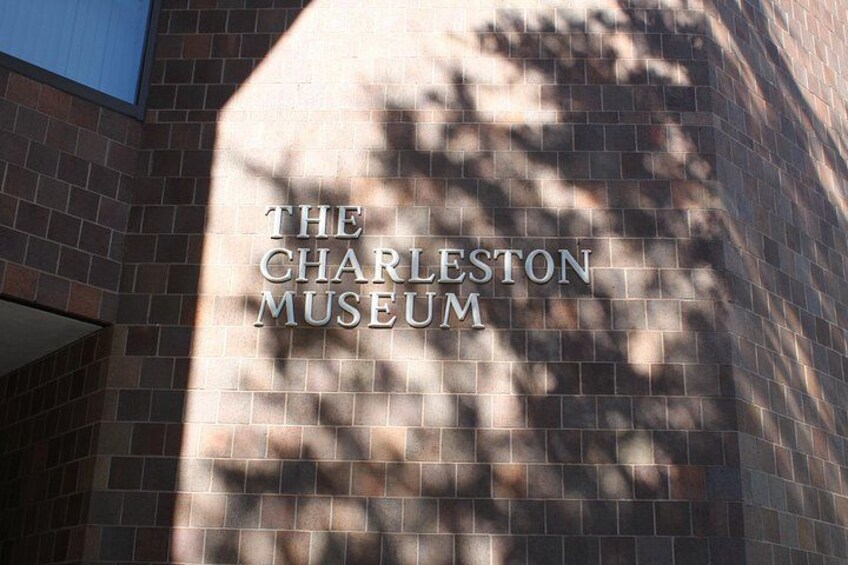 A brick wall with the text 'THE CHARLESTON MUSEUM' and shadows of trees. 1
