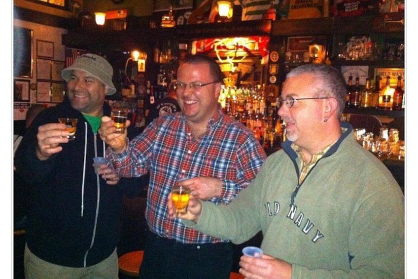 Three people holding drinks in a bar with shelves of bottles and a sign that reads 'No Navy'. 4
