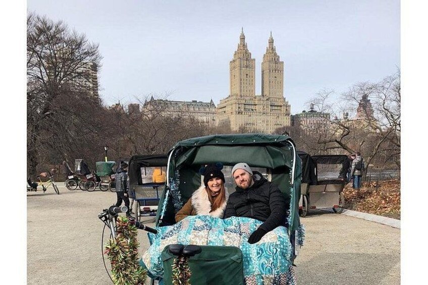 Un couple se trouve dans une voiture verte dont les sièges sont recouverts d’un tissu à motifs, à côté d’un vélo, et on aperçoit, au fond, un grand bâtiment doté de tours. 3