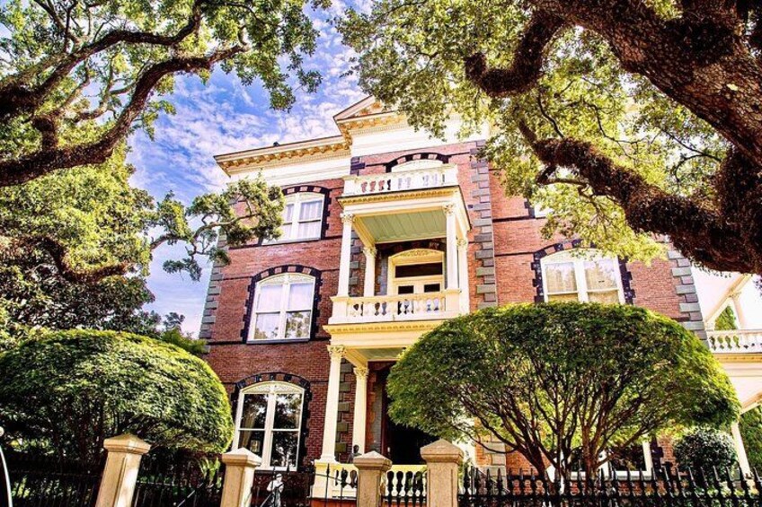 A Victorian-style house with a balcony, surrounded by trees and a wrought iron fence. 1