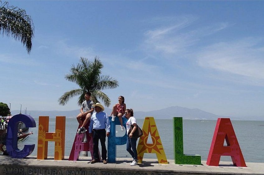 People standing in front of colorful letters spelling 'CHAPALA' by the water. 1