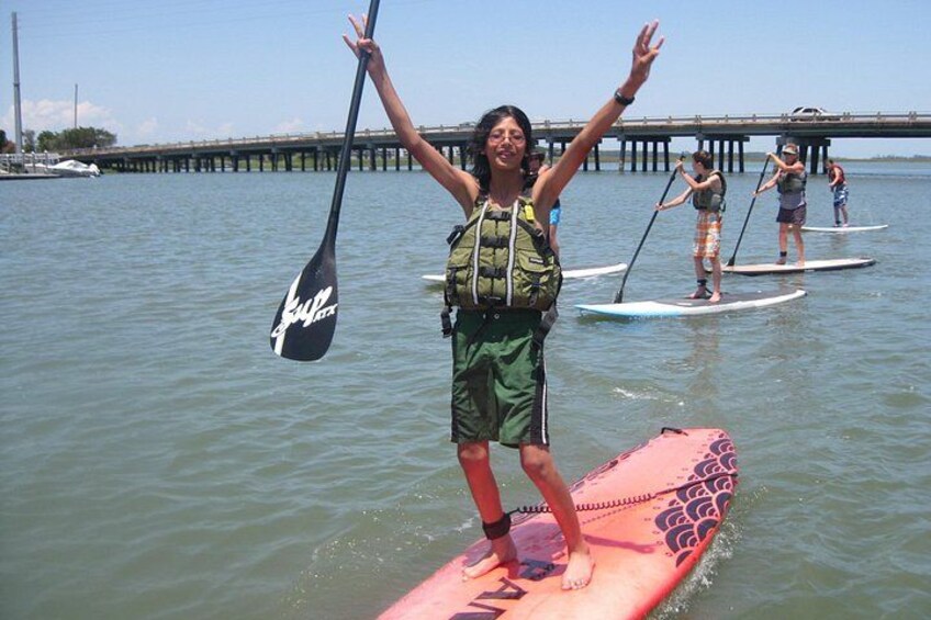 Une personne se tient debout sur une planche de paddle, les bras levés, une autre personne fait du paddle en arrière-plan, et une jetée s’avance dans l’eau. 3