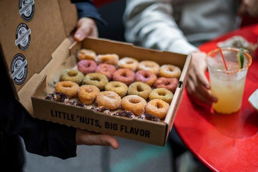 Una persona sostiene una caja de donas variadas, con una bebida al fondo. 3