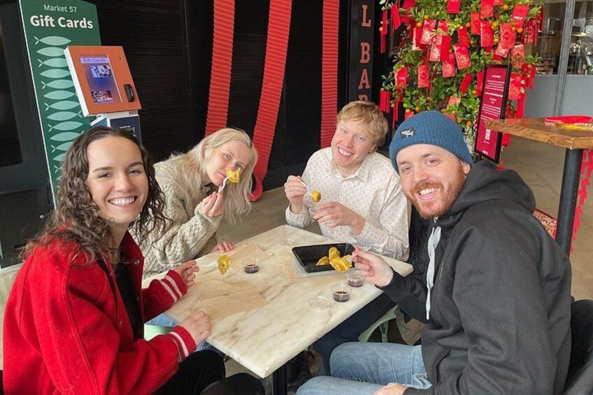 Four people sitting at a table with food and drink, a gift card machine, and a red decoration in the background. 3