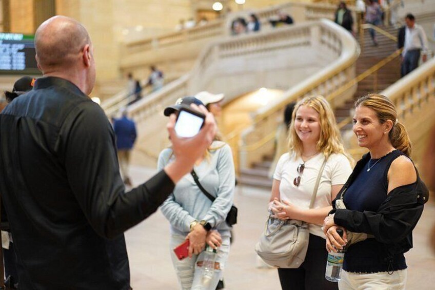 A man taking a photo of two women in a public indoor space with a staircase in the background. 2
