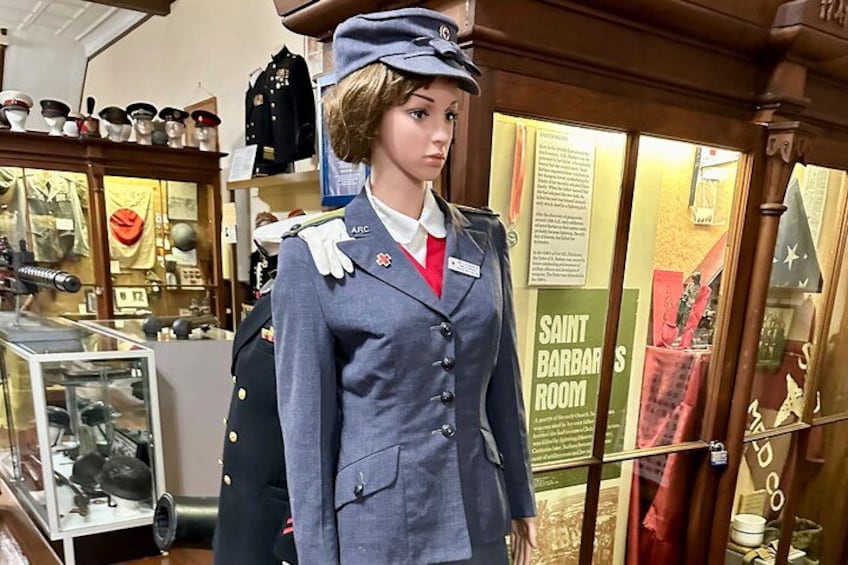 A mannequin dressed in a military uniform with a name tag, standing in a room with various military artifacts and a sign reading "SAINT BARBARS ROOM". 2