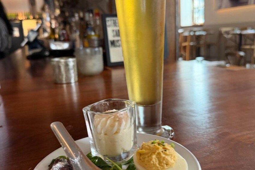 A bar setting with a glass of white wine, a shot glass with a creamy substance, and a plate with a green salad and a spoon. 1