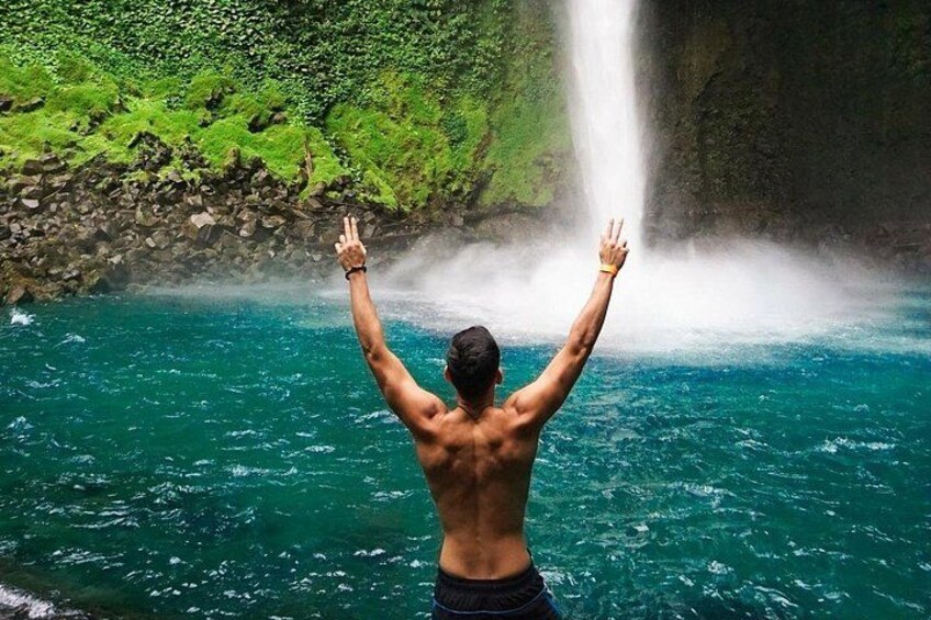 A person standing in water with arms raised, facing a waterfall. 1