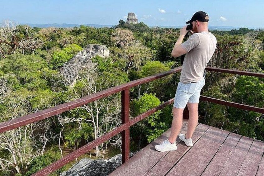 Eine Person fotografiert eine Maya-Pyramide von einer hölzernen Plattform mit Metallgeländer. 2