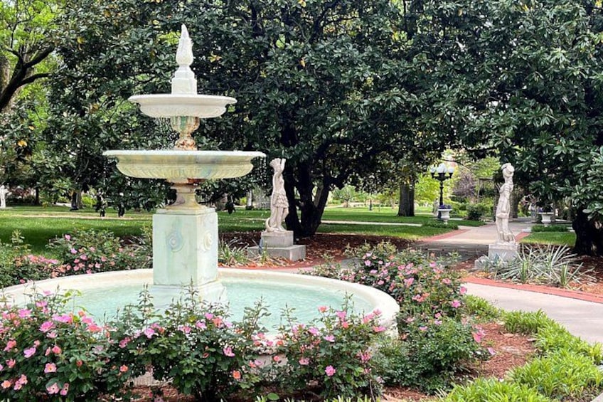 A multi-tiered fountain surrounded by pink flowers and greenery in a park with statues and trees. 2