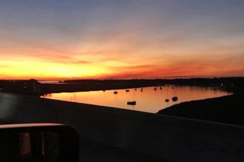 A sunset over a calm body of water with boats anchored, a bridge in the foreground, and a distant shoreline. 4