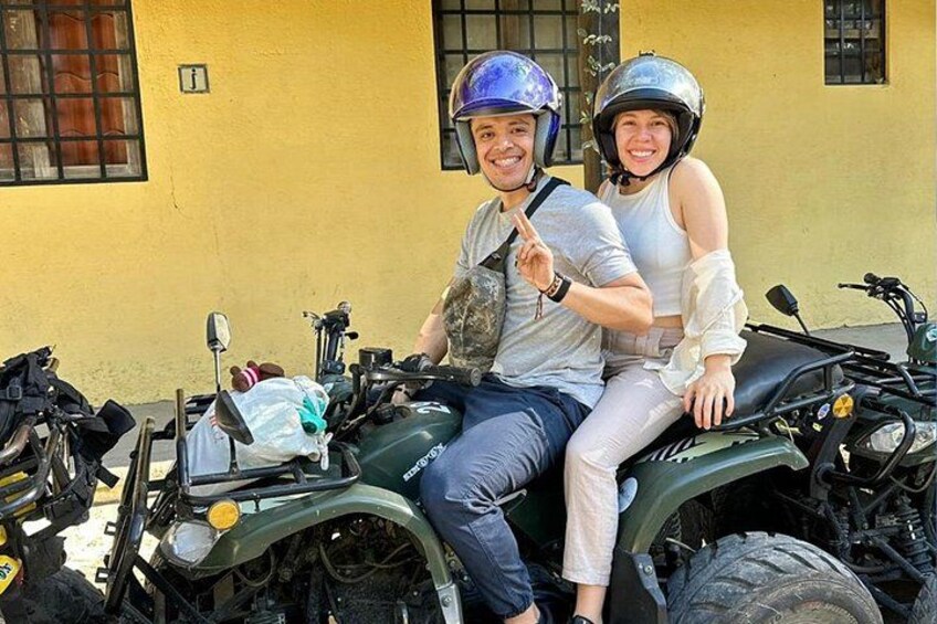 Deux personnes sur un quad vert, portant des casques, avec un bâtiment en arrière-plan et un sac sur le véhicule. 2