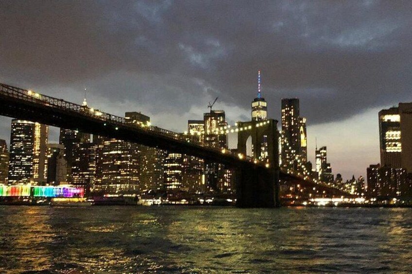 Une vue nocturne d’une ville, avec un pont imposant enjambant une rivière, des bâtiments illuminés et un ciel nuageux. 1