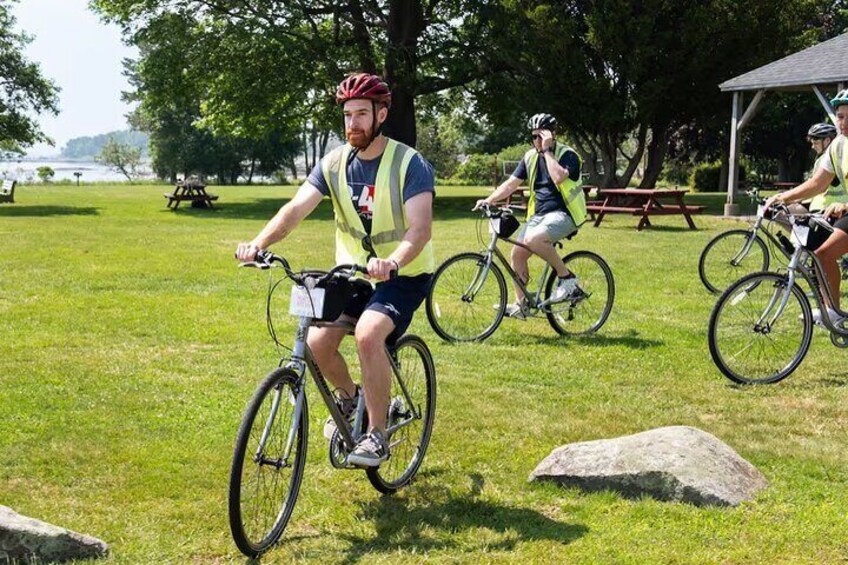 Three cyclists on a grassy path with picnic tables and a pavilion in the background. 1