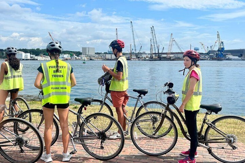 Four people wearing safety vests and helmets, standing by bicycles near a body of water with industrial structures in the background. 3
