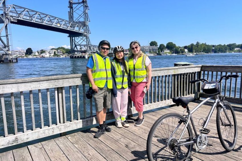 Trois personnes se trouvent sur une jetée en bois, avec un vélo, un pont et de l'eau en arrière-plan. 4