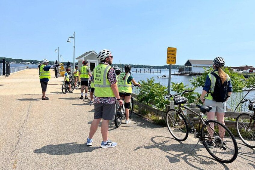 A group of cyclists on a bridge, wearing safety vests and helmets, with a body of water and buildings in the background. 3