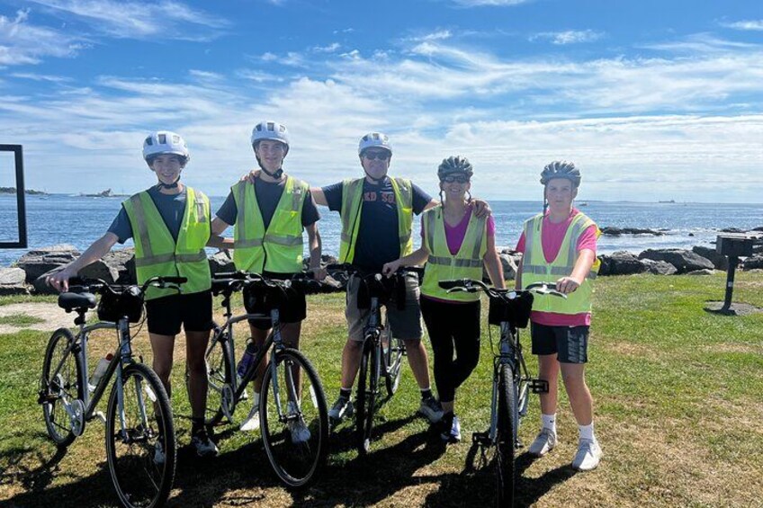 Four people on bicycles with safety vests, standing on grass near a body of water. 1