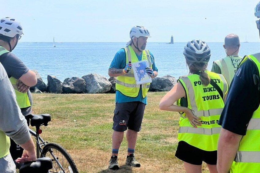 A group of people wearing safety vests and helmets, standing on grass near a body of water with rocks and a lighthouse in the background. 1