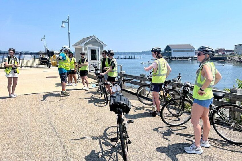 A group of people on a waterfront path with bicycles, wearing safety vests, and a small building in the background. 2