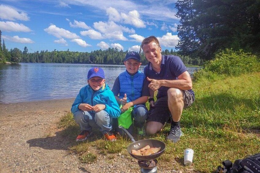 Tre personer sidder ved en sø med en pande mad og en dåse på jorden. 3