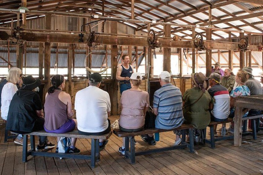 A group of people seated at wooden tables under a metal roof, listening to a speaker. 1