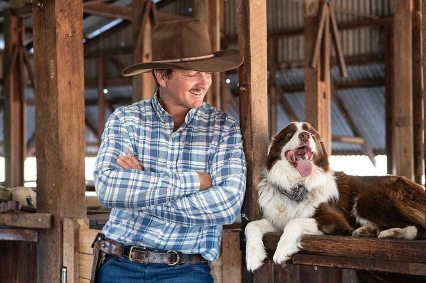 A person in a plaid shirt and cowboy hat standing next to a dog in a barn. 2