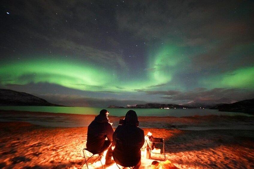 Two people sitting by a campfire under a night sky with the aurora borealis. 2