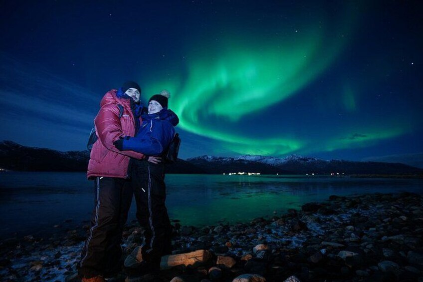 Two people standing on a rocky shore with a view of the northern lights over a body of water. 3