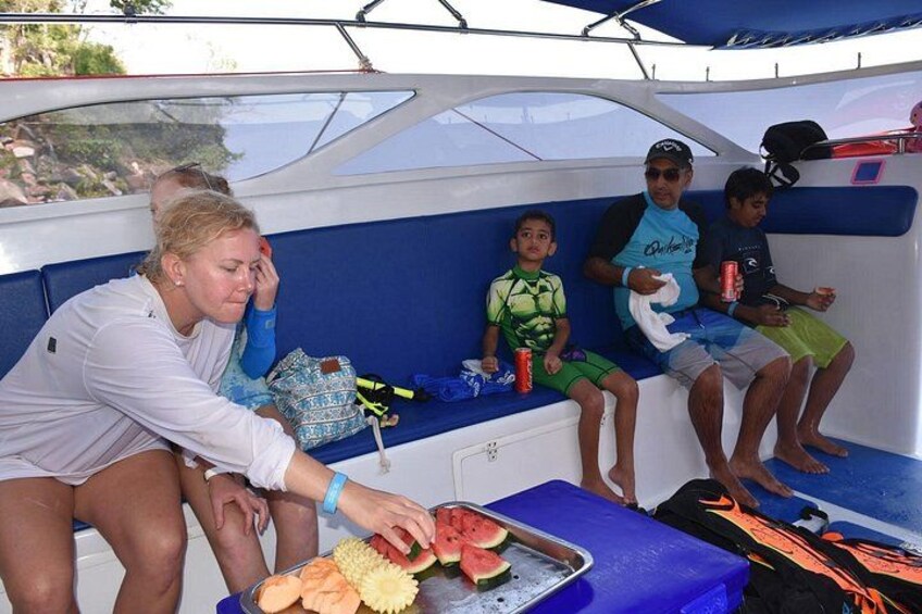 A boat with people sitting on the deck, a watermelon, and a tray of food. 2