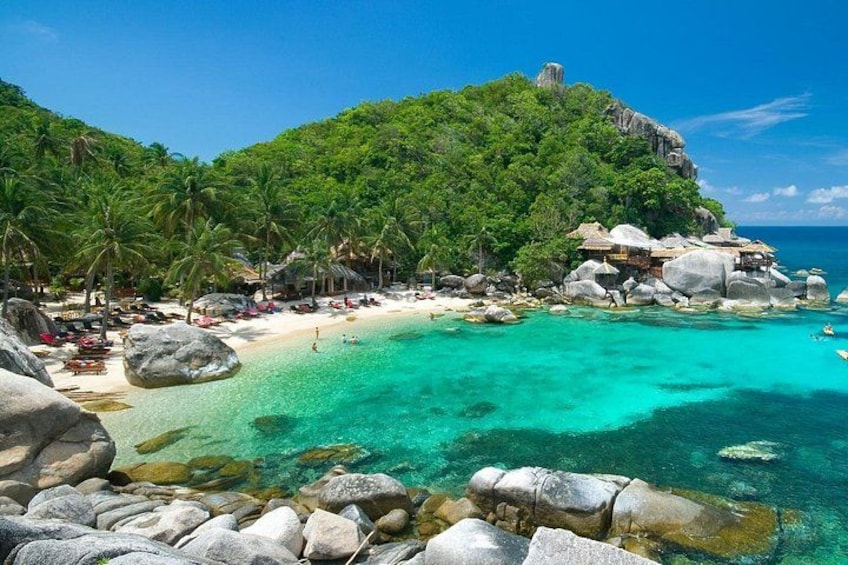 A tropical beach with clear turquoise water, large rocks, palm trees, and a small hill with a structure on top. 1