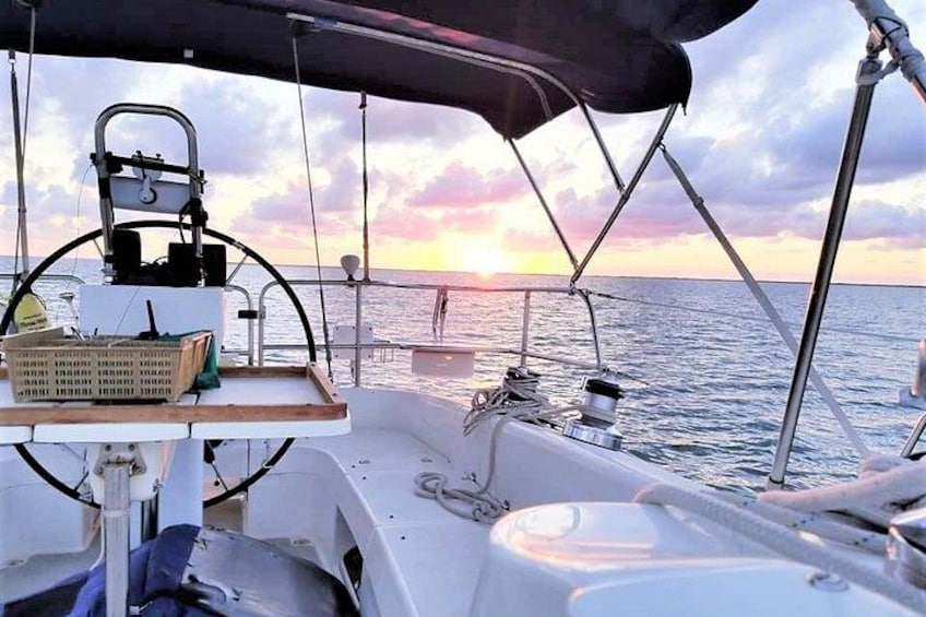 Het dek van een zeilboot met een stuurrad, een mand en een zonsondergang aan de horizon. 1